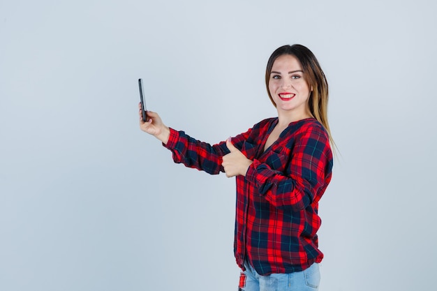 Jeune belle femme en chemise décontractée prenant selfie, montrant le pouce vers le haut et à la félicité, vue de face.