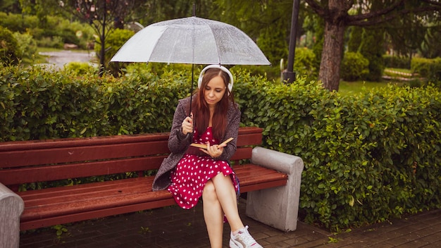 Jeune belle femme dans des écouteurs avec un parapluie transparent lisant un livre assis sur un banc dans le parc de la ville sous la pluie