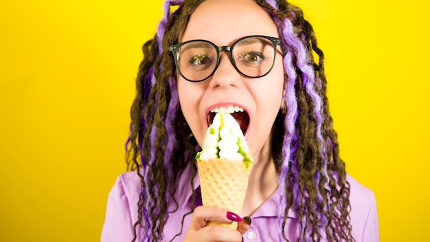 Jeune belle femme dans des verres mordre la crème glacée en regardant la caméra en souriant Jolie femme brillante avec des dreadlocks en chemise violette, manger des coupes glacées sur fond jaune en studio Ralenti