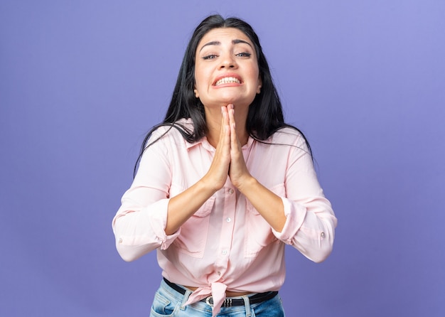 Jeune belle femme dans des vêtements décontractés tenant les mains ensemble comme prier avec l'expression d'espoir mendier debout sur le mur bleu