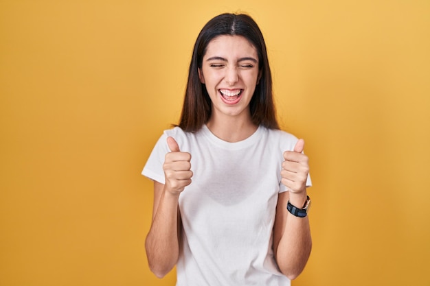 Jeune belle femme debout sur fond jaune excitée pour le succès avec les bras levés et les yeux fermés célébrant la victoire en souriant. notion de gagnant.