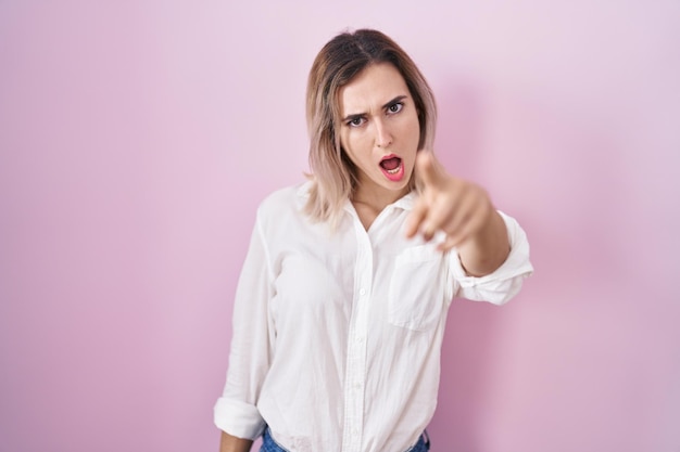 Jeune belle femme debout sur fond rose pointant mécontent et frustré vers la caméra, en colère et furieuse contre vous