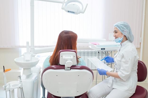 Jeune belle femme dentiste parlant à un patient au bureau Une femme rousse est assise sur une chaise avec un orthodontiste et écoute les rendez-vous Services dentaires Vue de l'arrière