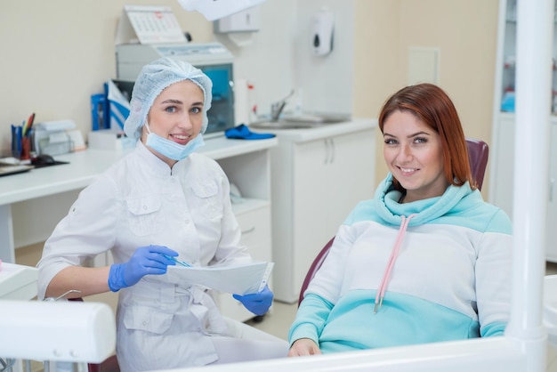 Jeune belle femme dentiste et patiente heureuse regardent directement dans la caméra Une femme rousse est assise sur une chaise avec un orthodontiste et sourit Services dentaires