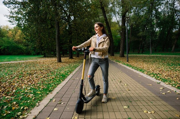 Jeune belle femme élégante équitation scooter électrique par parc d'automne, transport écologique