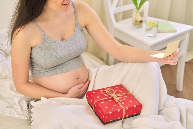 Jeune belle femme enceinte avec boîte-cadeau, femme assise à la maison dans son lit en lisant une carte de voeux