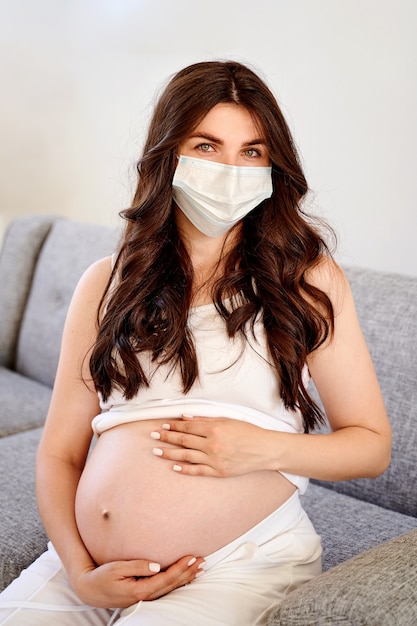 Jeune belle femme enceinte en masque chirurgical médical et t-shirt blanc mis en quarantaine à la maison par la fenêtre. Coronavirus, virus.