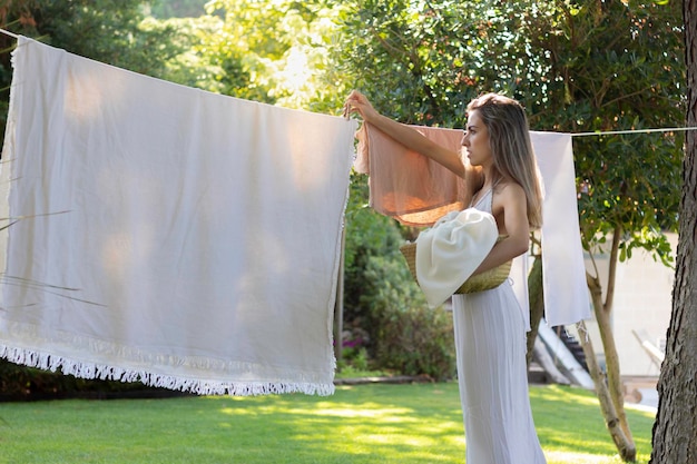 Jeune et belle femme étendant le linge dans le jardin en robe blanche et entourée par la nature