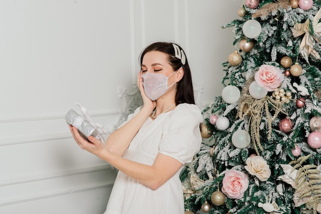 Jeune belle femme européenne en masque médical avec des cadeaux de vacances à la maison, coronavirus de quarantaine.