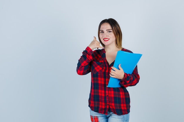 Jeune belle femme faisant un geste téléphonique, tenant un dossier en chemise décontractée, un jean et l'air gai. vue de face.