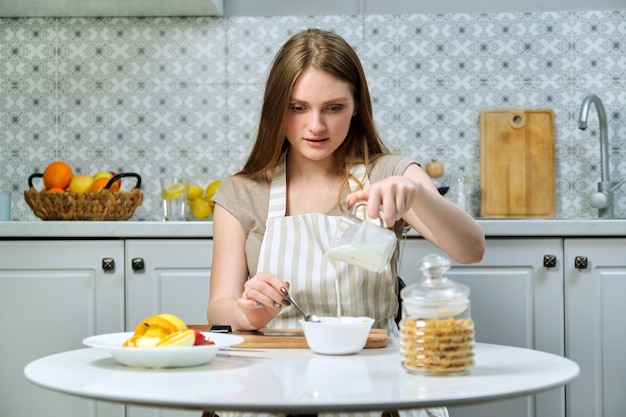Jeune belle femme faisant une salade de fruits dans la cuisine avec pomme, orange, yaourt, flocons de maïs et graines. Alimentation saine cuisinée à la maison, beauté et santé des jeunes