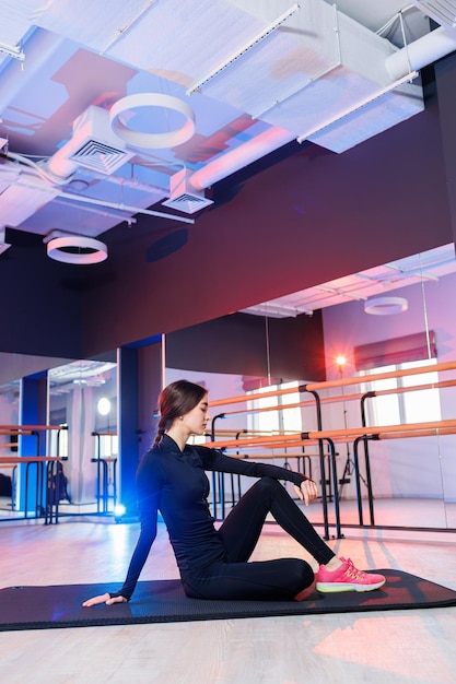 Jeune belle femme flexible en costume noir est assise sur le sol et travaille dur sur elle-même et s'étire