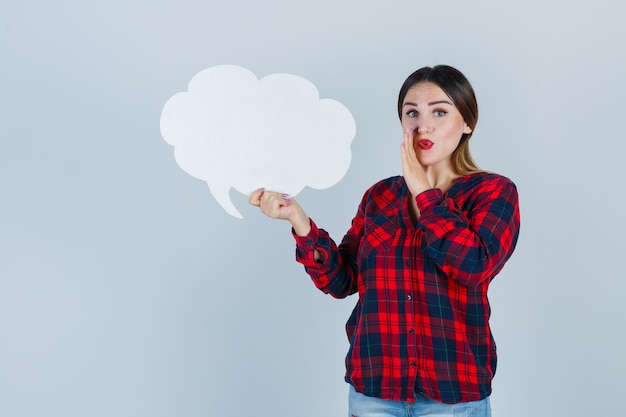 Jeune belle femme gardant la main près de la bouche tout en tenant une bulle de dialogue en chemise décontractée, un jean et l'air concentré, vue de face.