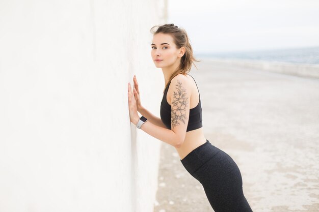 Jeune belle femme en haut sportif gris foncé et leggings regardant rêveusement à huis clos tout en s'appuyant sur le mur faisant des exercices en plein air