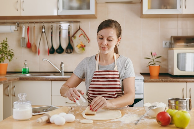 La jeune belle femme heureuse assise à une table avec de la farine et découpe un verre dans un cercle de pâte pour les boulettes dans la cuisine. Cuisiner à la maison. Préparer la nourriture.