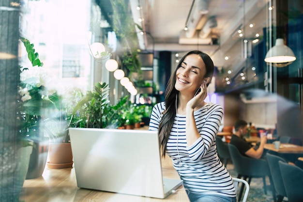 Jeune belle femme heureuse dans les écouteurs écoute de la musique sur un ordinateur portable dans un café ou un centre de travail