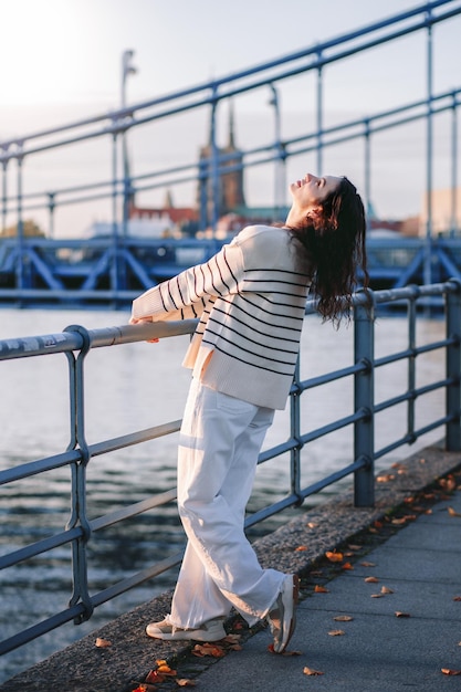 Jeune belle femme hipster souriante dans des vêtements d'été à la mode Modèle insouciant sexy posant sur le fond de la rue d'europe au coucher du soleil Femme brune positive Enthousiaste et heureuse