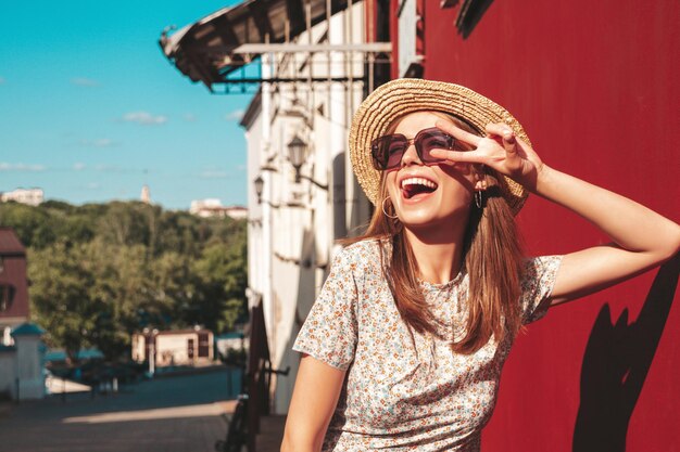 Jeune belle femme hipster souriante en robe d'été à la mode Sexy femme insouciante posant sur le fond de la rue près du mur rouge en chapeau au coucher du soleil Modèle positif à l'extérieur Enthousiaste et heureux