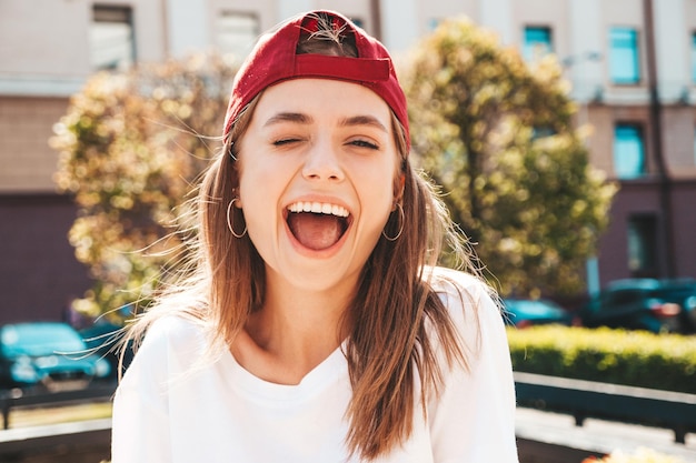 Jeune belle femme hipster souriante en t-shirt blanc d'été à la mode Sexy femme insouciante posant sur le fond de la rue en casquette au coucher du soleil Modèle positif à l'extérieur Enthousiaste et heureux