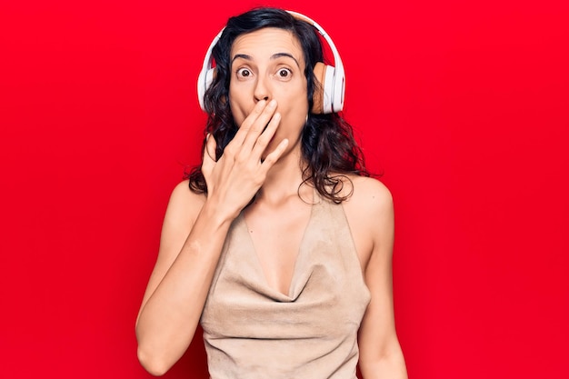 Jeune belle femme hispanique écoutant de la musique à l'aide d'un casque couvrant la bouche avec la main choquée et effrayée par une expression surprise d'erreur