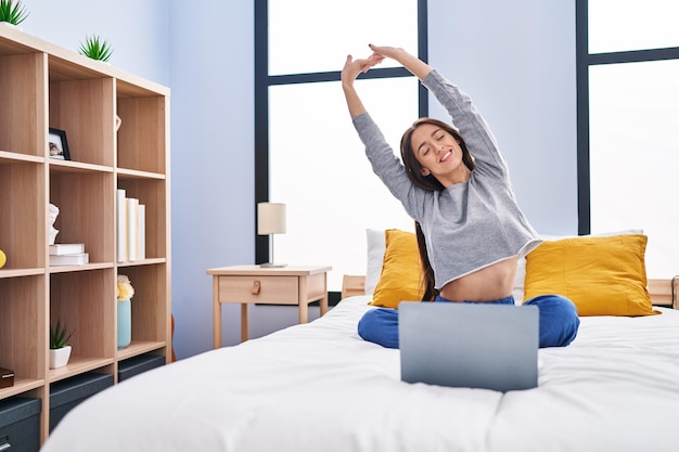 Jeune belle femme hispanique faisant des exercices d'étirement en ligne assis sur le lit dans la chambre