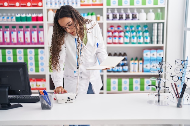 Jeune belle femme hispanique pharmacien parlant au téléphone lisant un cahier à la pharmacie