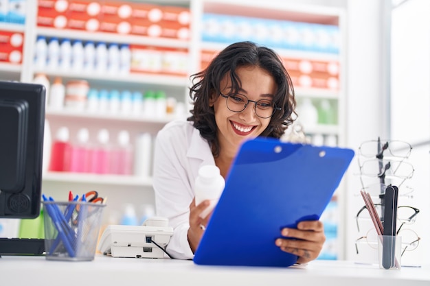 Jeune belle femme hispanique pharmacien tenant une bouteille de pilules lisant une ordonnance à la pharmacie