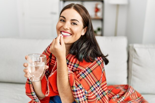 Une jeune et belle femme hispanique qui prend des pilules assise sur le canapé à la maison.