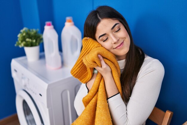 Jeune belle femme hispanique souriante confiante touchant des vêtements adoucissants avec le visage à la buanderie