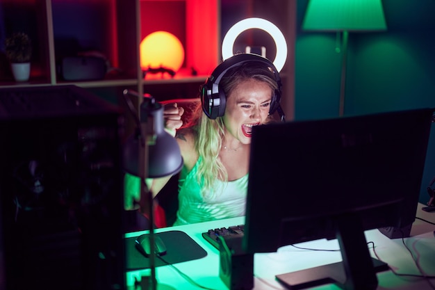 Jeune belle femme hispanique streamer jouant au jeu vidéo avec l'expression du gagnant dans la salle de jeux