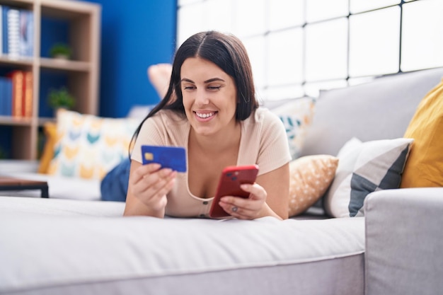 Jeune belle femme hispanique utilisant un smartphone et une carte de crédit allongée sur un canapé à la maison