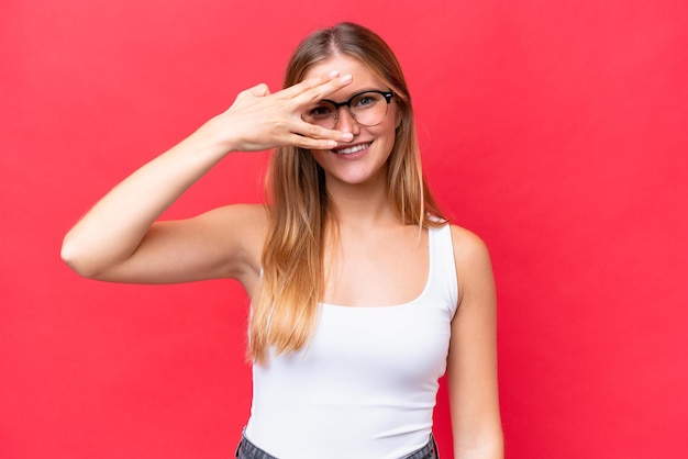 Jeune belle femme isolée sur fond rouge couvrant les yeux par les mains et souriant