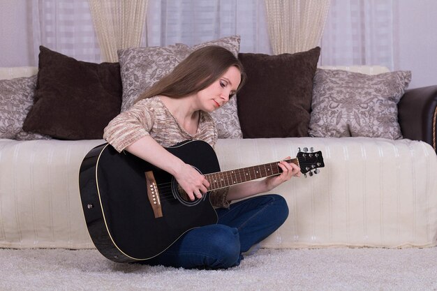 Jeune belle femme jouant de la guitare noire assise sur un tapis à la maison