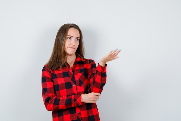 Jeune belle femme levant la main de manière interrogative, regardant loin en chemise décontractée et l'air perplexe. vue de face.