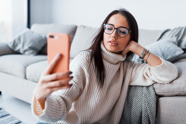 Jeune belle femme à lunettes assise seule à la maison avec un téléphone dans les mains.