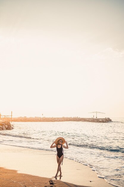 Jeune belle femme en maillot de bain noir et chapeau à lunettes se promène le long de la plage en Turquie au coucher du soleil Le concept de loisirs en mer Mise au point sélective