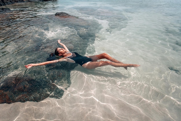 Jeune belle femme en maillot de bain noir se détend et prend un bain de soleil allongé dans l'eau sur la plage. La vue du haut