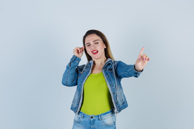 Jeune Belle Femme Avec La Main Près Du Visage Tout En Pointant Vers Le Haut En Tenue De Denim Et L'air Gaie, Vue De Face.