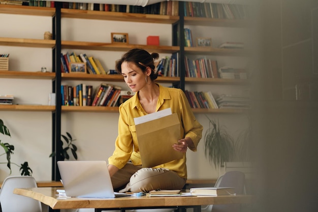 Jeune belle femme pensive en chemise jaune assise sur un bureau avec des papiers tout en travaillant rêveusement sur un ordinateur portable dans un bureau moderne