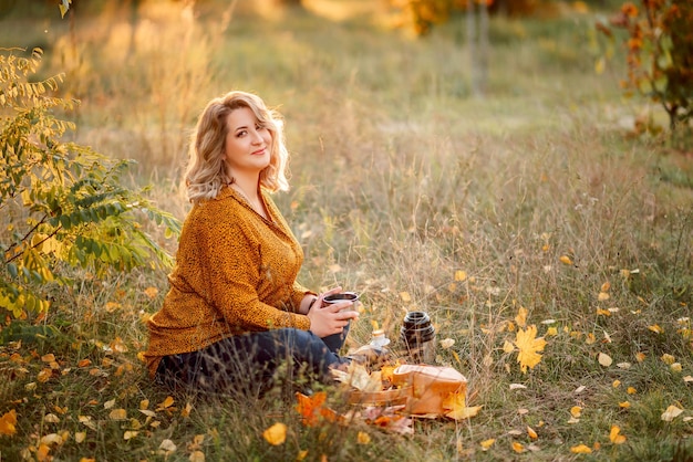 Jeune belle femme plussize dans une chemise orange et un jean est assise dans un parc d'automne Boire du thé et se détendre sur un pique-nique