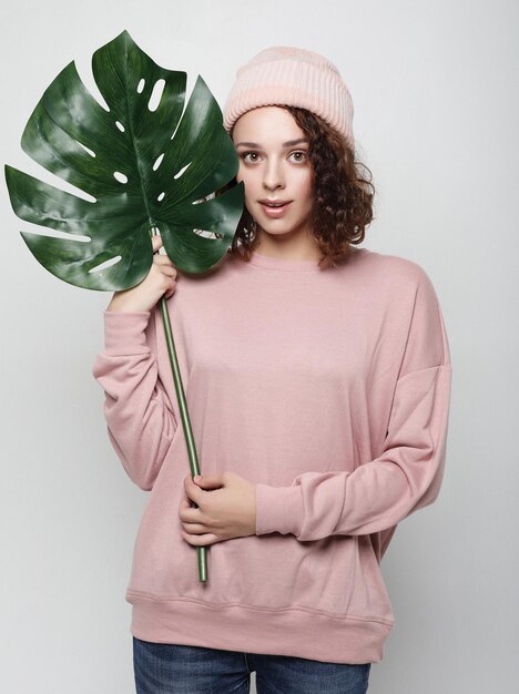 Jeune belle femme portant des vêtements décontractés roses tenant une feuille de monstera posant sur fond blanc