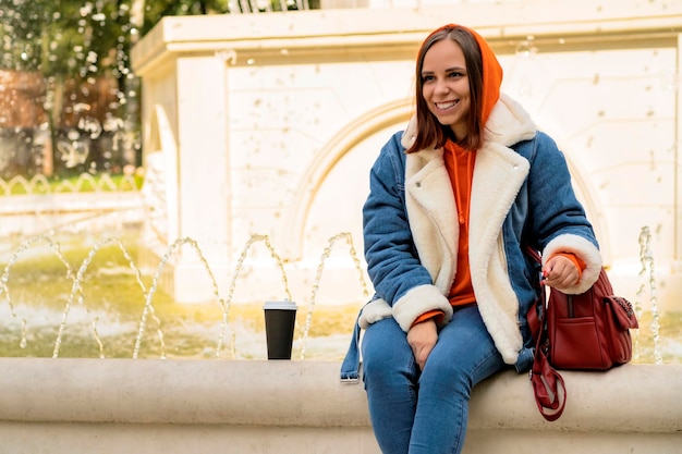 Jeune belle femme positive dans des vêtements chauds décontractés avec du café dans une tasse en papier assis sur une fontaine dans le parc de la ville