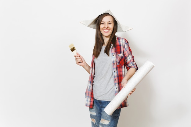 Jeune belle femme ravie dans des vêtements décontractés et un chapeau de journal tenant un pinceau et un rouleau de papier peint isolé sur fond blanc. Instruments pour la salle d'appartement de rénovation. Réparer le concept de maison.