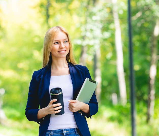 Jeune belle femme souriante dans le parc avec un gadget de tablette et un casque sans fil profitant du travail à distance du concept d'énergie verte