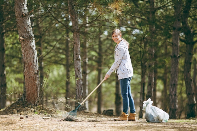 Jeune belle femme souriante nettoyant et utilisant un râteau pour la collecte des ordures près des sacs poubelles dans un parc ou une forêt. Problème de pollution de l'environnement