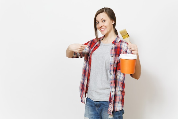 Jeune belle femme souriante pointant l'index sur le pinceau et le seau de peinture isolé sur fond blanc. Instruments, accessoires, outils pour la salle d'appartement de rénovation. Réparer le concept de maison.