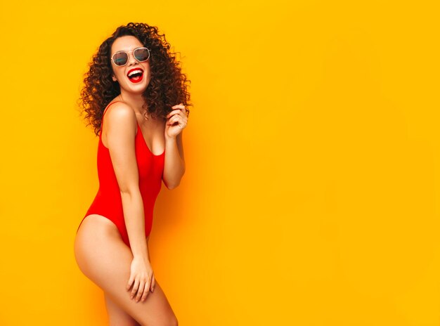 Jeune belle femme souriante posant près du mur jaune en studioModèle sexy en maillot de bain rouge