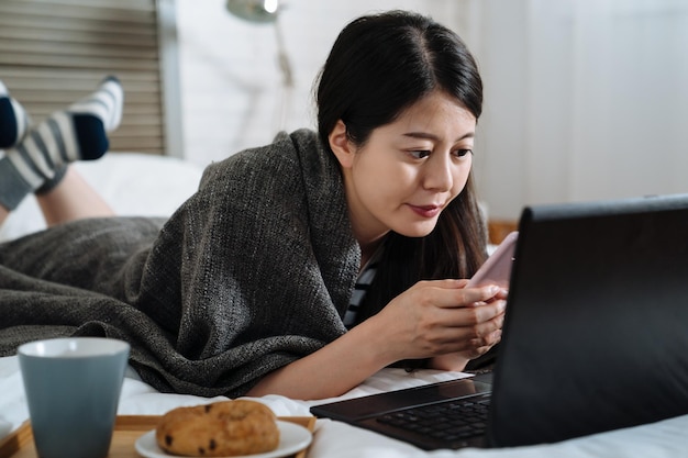 Jeune belle femme souriante prenant son petit déjeuner au lit et utilisant un téléphone portable technologique. une fille coréenne asiatique aime manger du pain et boire une tasse de café sur un plateau dans un endroit confortable blanc couché couverture couverture