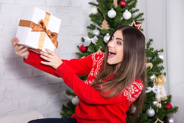 Jeune belle femme surprise ouvrant une boîte-cadeau près d'un arbre de Noël décoré