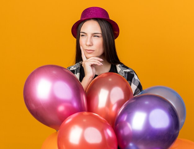 Jeune belle femme suspecte portant un chapeau de fête debout derrière des ballons mettant la main sur la joue isolée sur un mur orange